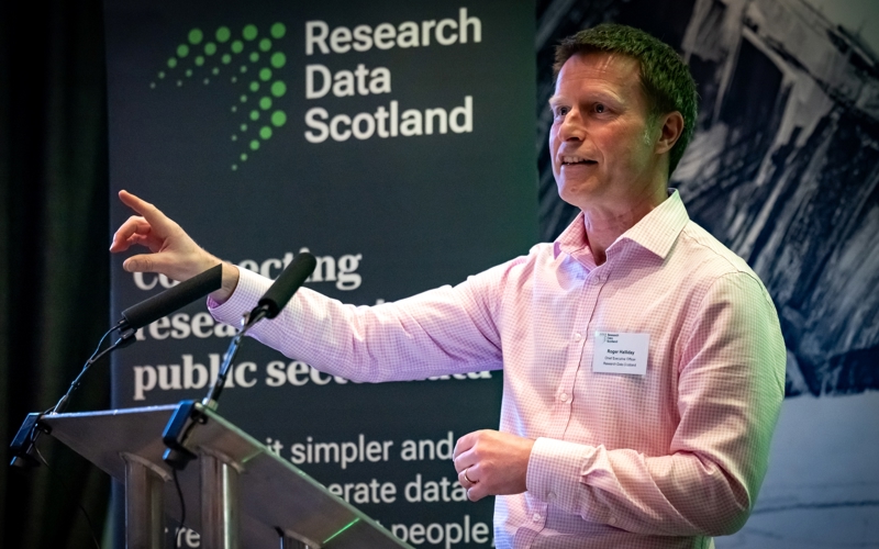 A man giving a speech at a metal lectern. A banner in the background displays the Research Data Scotland logo.