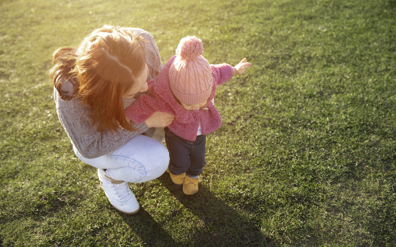 A woman in grey jumper and jeans bends down to support a walking toddler dressed in pink. 