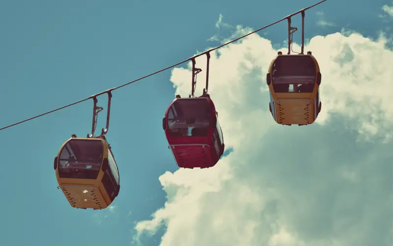 Three cable cars, two yellow and one red, hang against a backdrop of blue sky and white clouds. 
