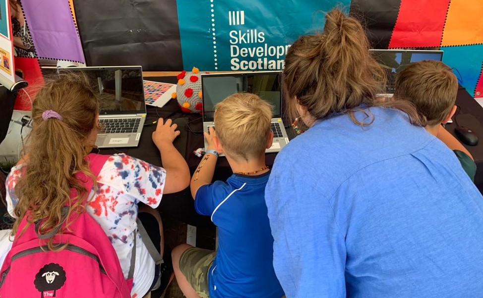 Three children sit at laptops, facing away from the camera, playing Minecraft