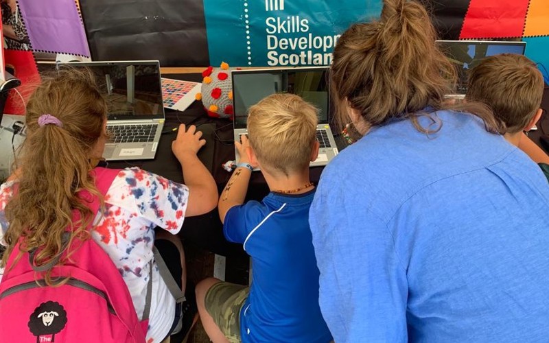 Three children sit at laptops, facing away from the camera, playing Minecraft