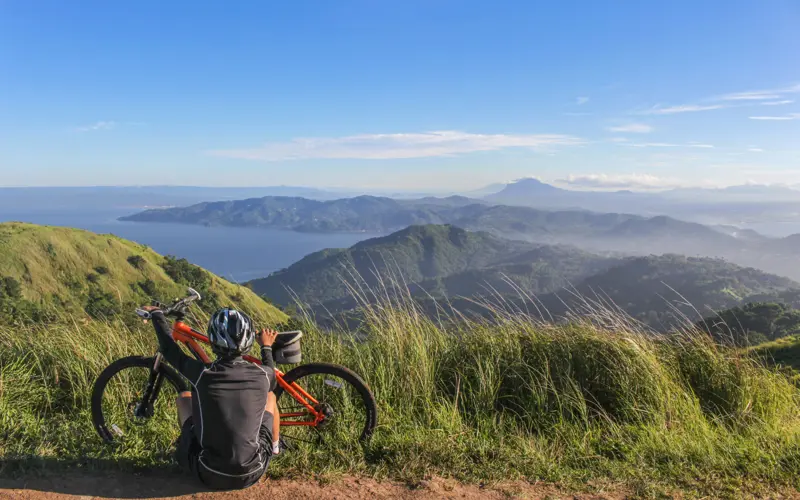 A cyclist sits atop a mountain range, looking out at the scenery with their back to the camera.