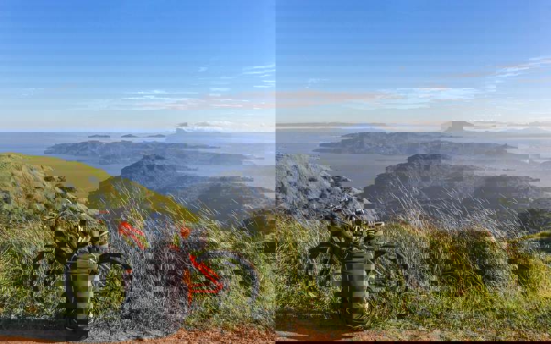 A cyclist sits atop a mountain range, looking out at the scenery with their back to the camera.