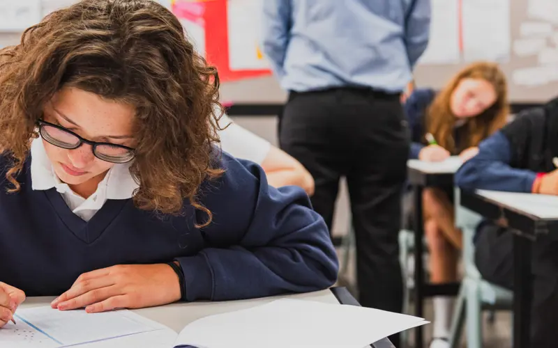 Young people at desks bow their heads over exam papers. 