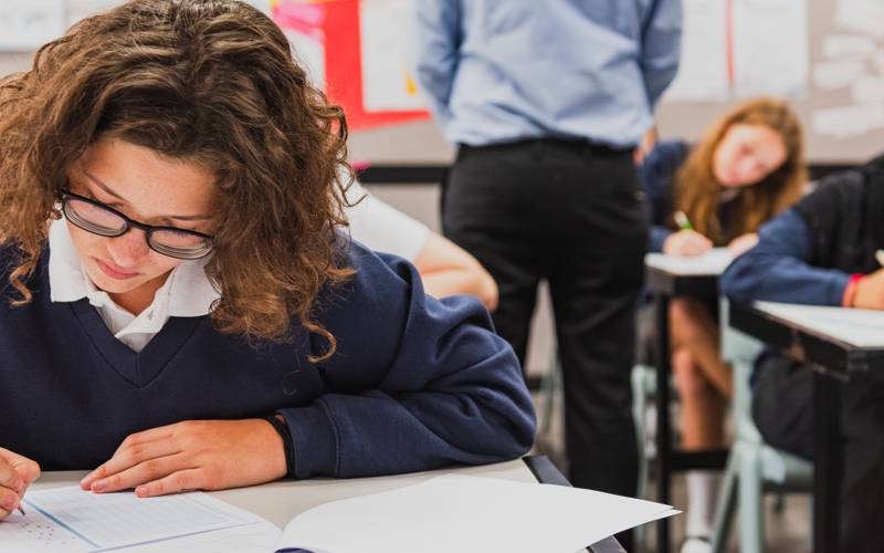 Young people at desks bow their heads over exam papers. 
