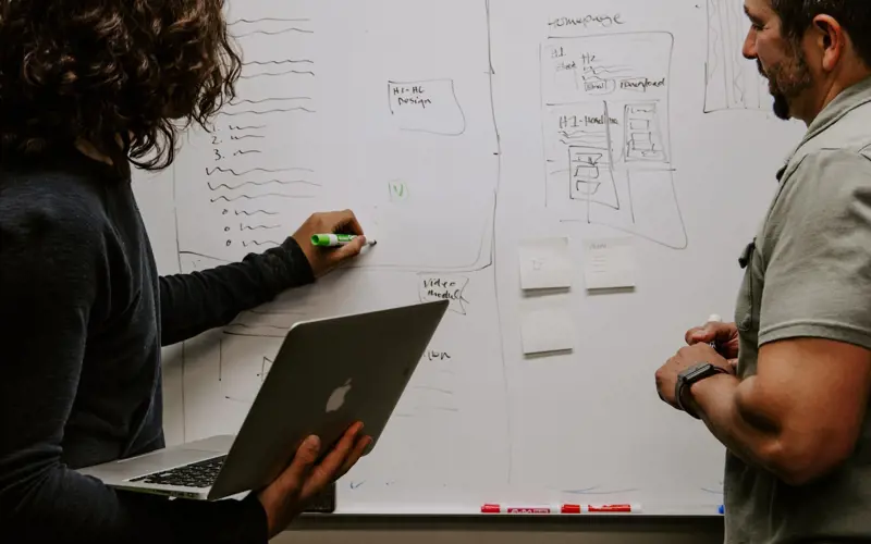 A person holding a laptop writing on a whiteboard as another person looks on