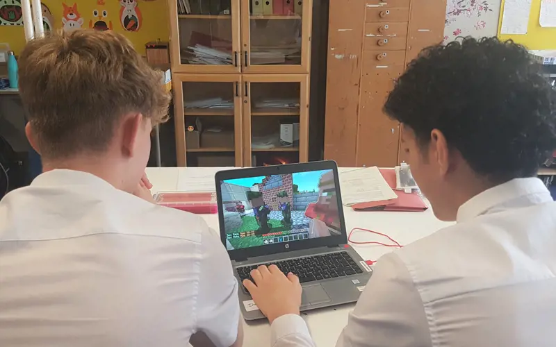 Two teenage boys sit, facing away from the camera and looking at a laptop running the game Minecraft