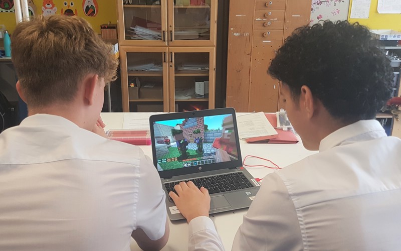 Two teenage boys sit, facing away from the camera and looking at a laptop running the game Minecraft