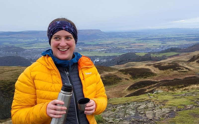 Rosie Seaman stands on top of a hill, holding a flask and smiling