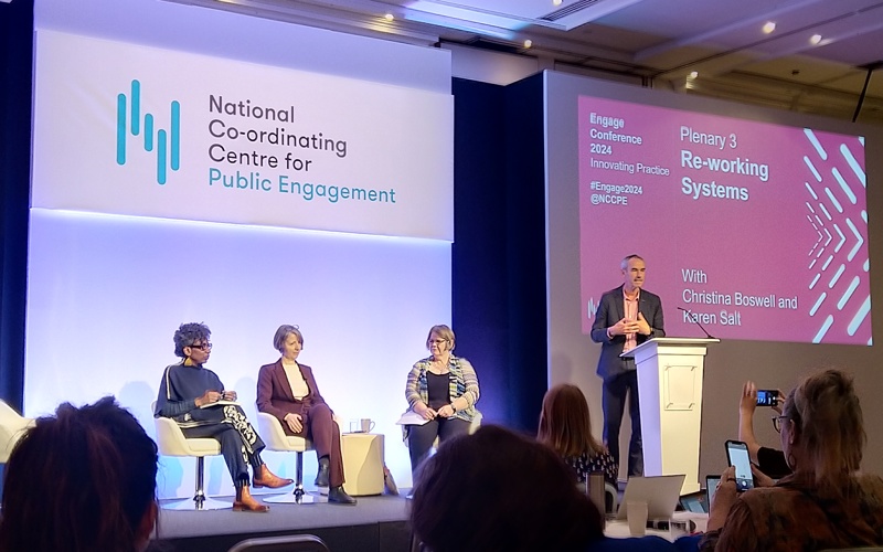 Four people on a stage. One of them is presenting to a room of people.