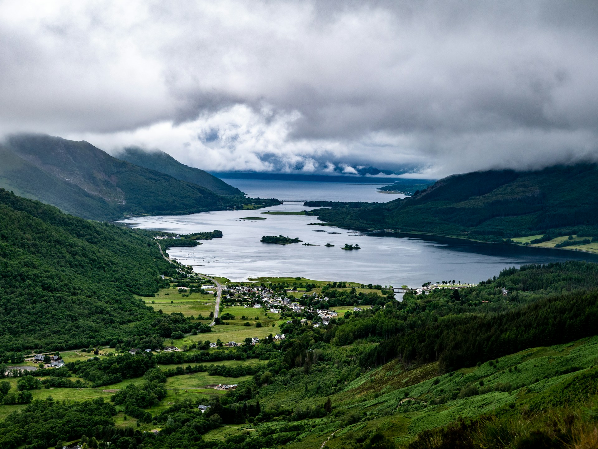 Photo of Loch Leven. A green valley covered in trees, with buildings at the base and a large loch spreading to the horizon.