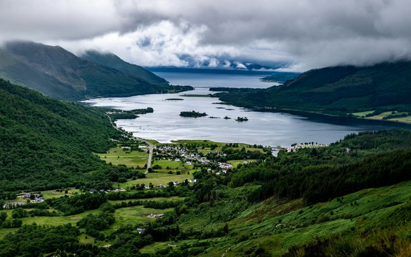 Photo of Loch Leven. A green valley covered in trees, with buildings at the base and a large loch spreading to the horizon.