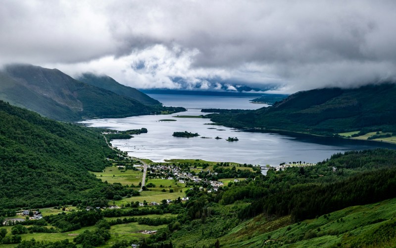 Photo of Loch Leven. A green valley covered in trees, with buildings at the base and a large loch spreading to the horizon.