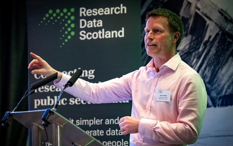 A man giving a speech at a metal lectern. A banner in the background displays the Research Data Scotland logo.