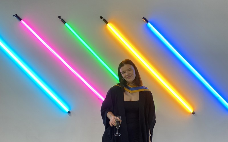 Stella Telford in graduation robes stands in front of multi-coloured lighting installation