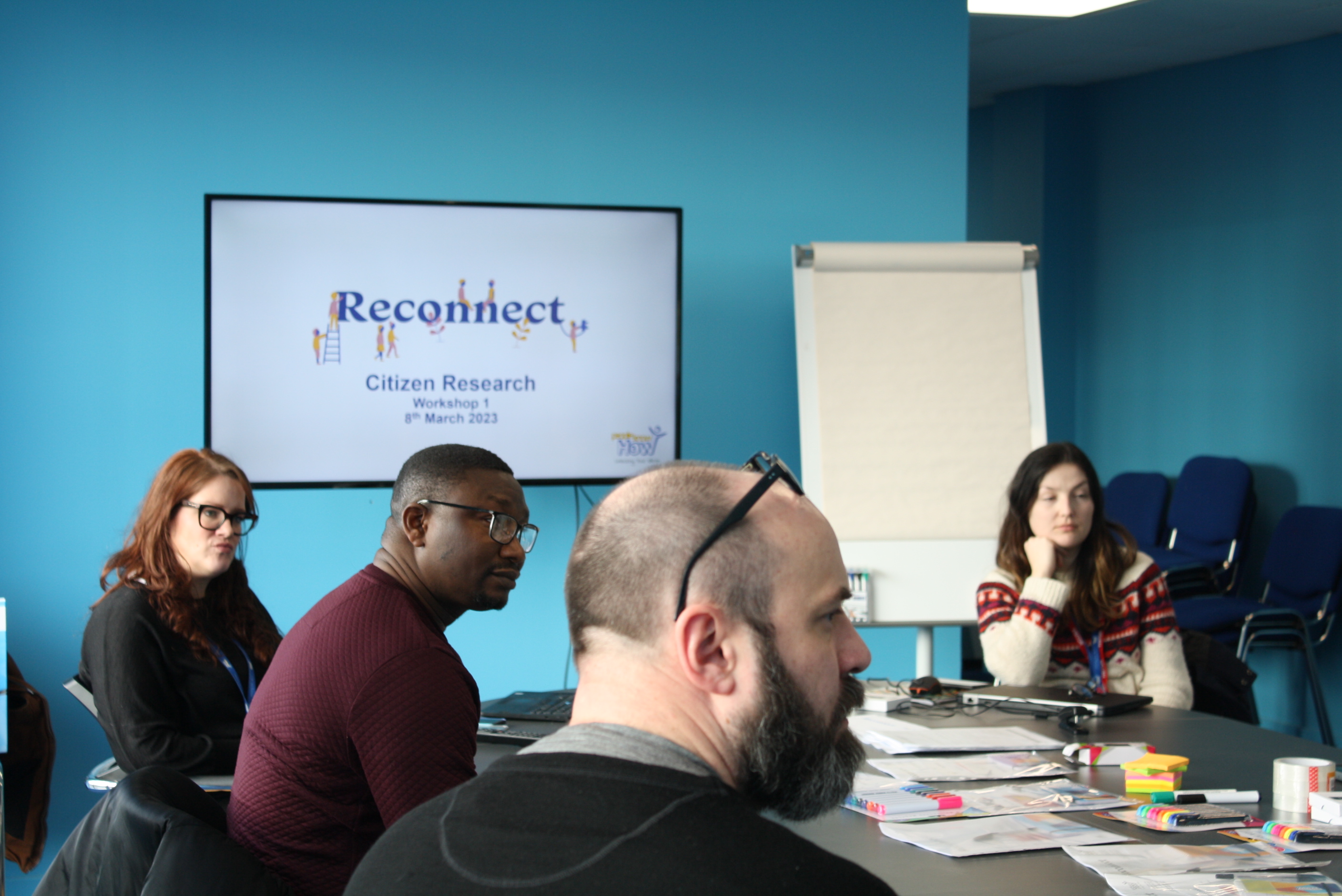 A group of people sit around a table looking at someone off-camera. A screen in the background displays the words 