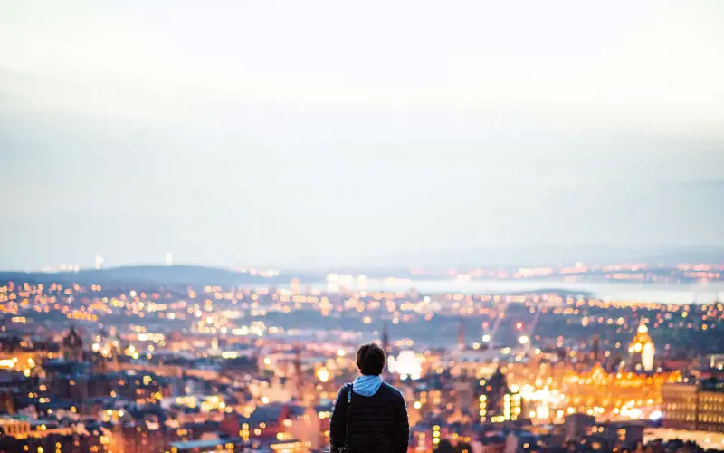 A person in the foreground looks out on the lights of Edinburgh in the evening.