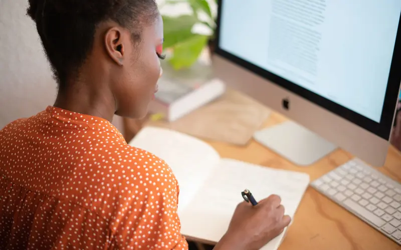 A woman sits in front of a desktop computer writing in a notebook.