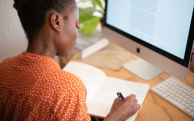 A woman sits in front of a desktop computer writing in a notebook.