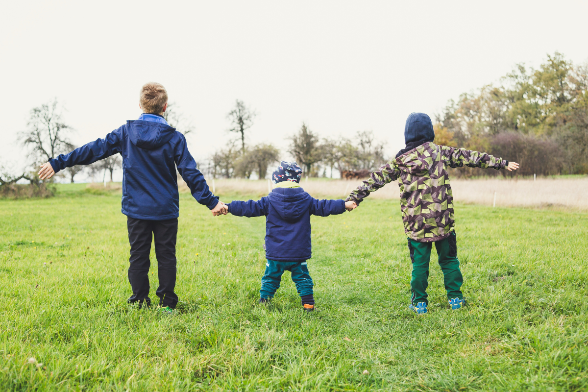 Three children hold hands standing in grass, with their backs towards the camera