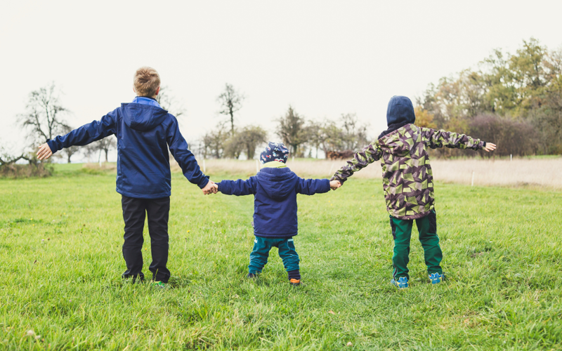 Three children hold hands standing in grass, with their backs towards the camera