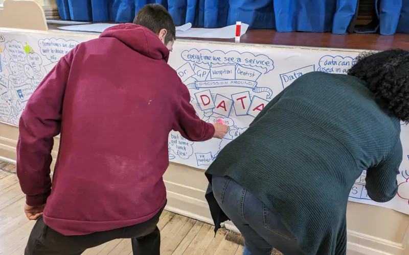 Two people facing away from the camera are sticking post-it notes onto a long sheet of paper. The paper has notes from a discussion about data written and illustrated on it