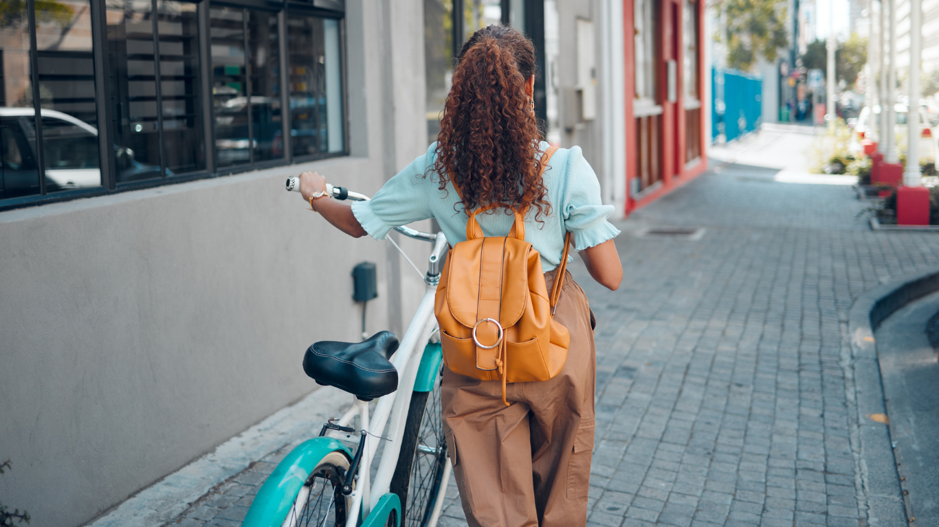 Woman  with bicycle and backpack faces away from the camera as she makes her way down a cobbled street.