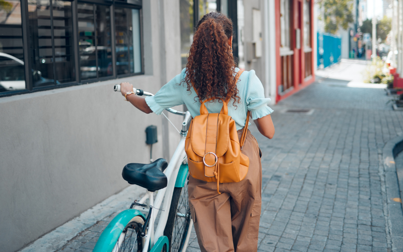 Woman  with bicycle and backpack faces away from the camera as she makes her way down a cobbled street.