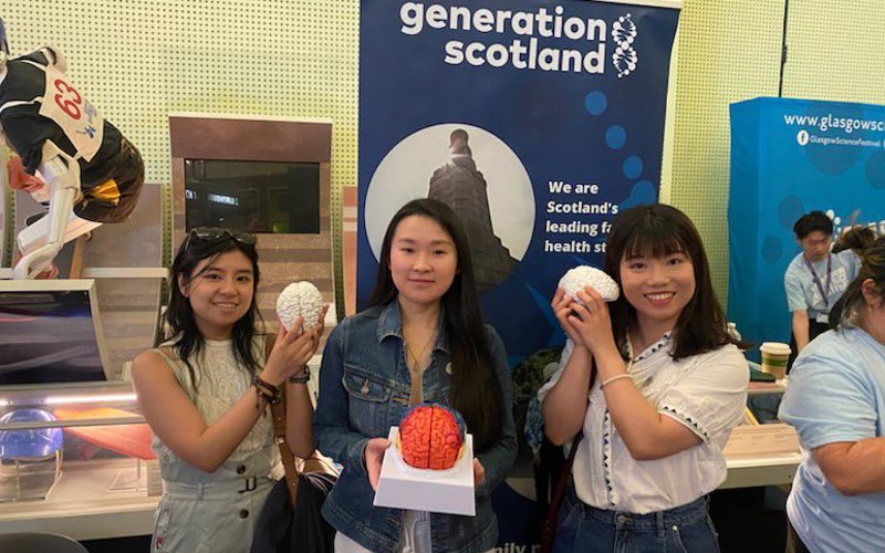 Three people hold models of brains. They are standing in front of a banner that reads 