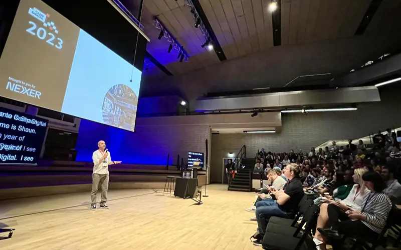 A person stands in a large room, speaking towards a crowd. A screen above him has the words 