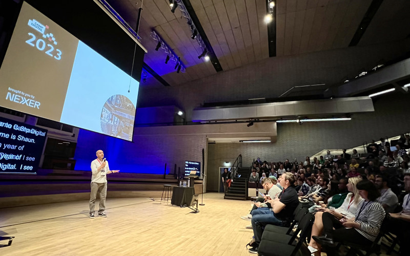 A person stands in a large room, speaking towards a crowd. A screen above him has the words 