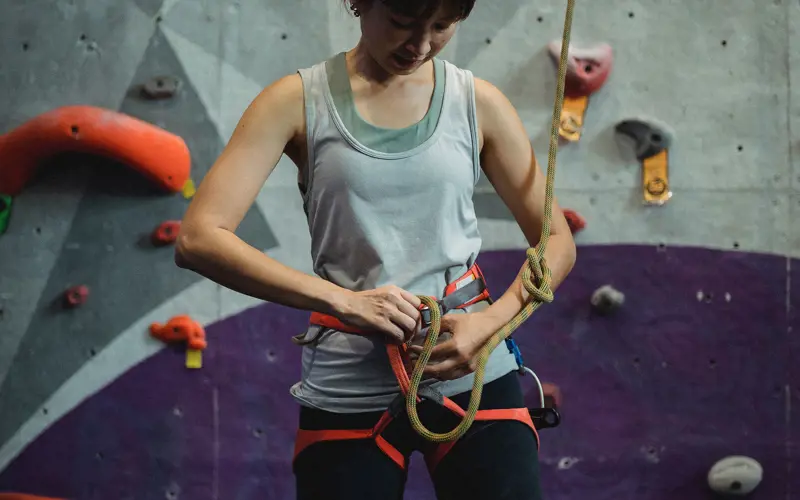 A woman in front of a climbing wall fastens a climbing rope to the harness on her waist.
