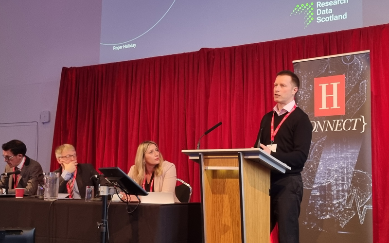 Roger Halliday stands at a podium and presents to an audience, with panel members in frame to the left. 