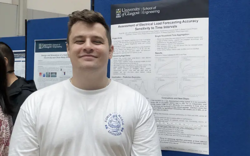 A young white man, Liam McAlister, stands smiling in front of an academic poster